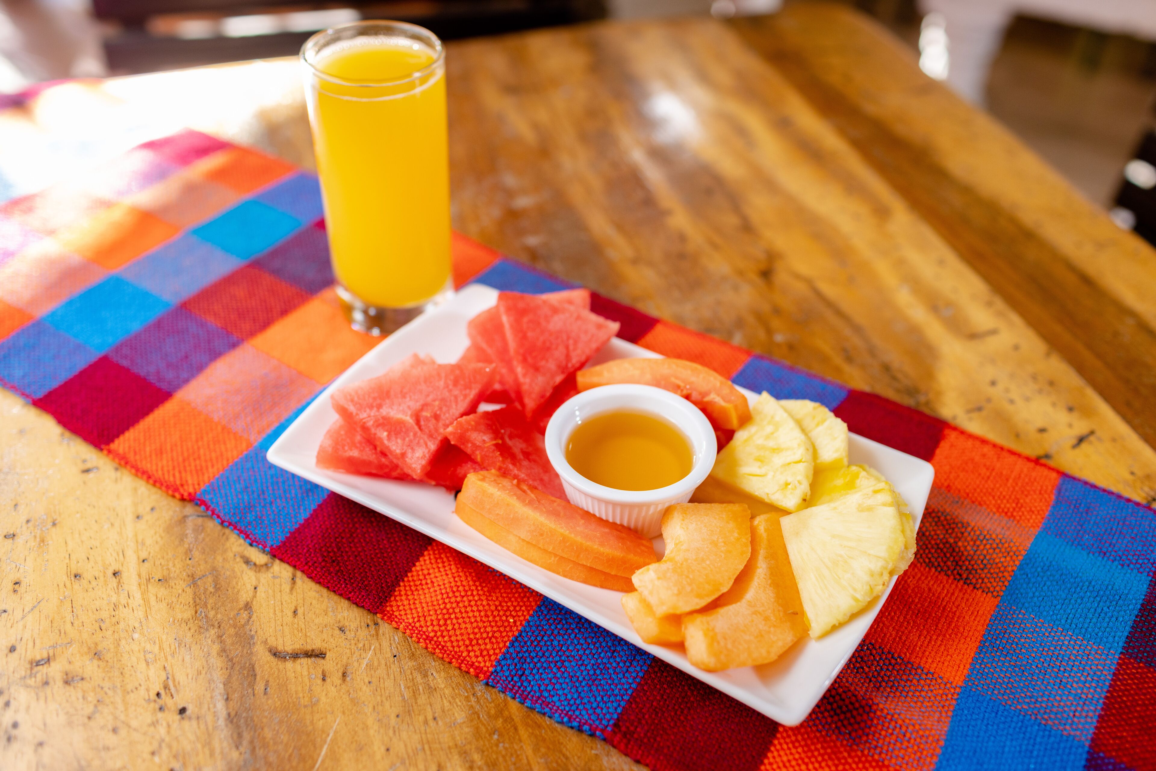 Plato con sandía, piña y papas fritas, acompañado de un vaso de jugo de naranja, sobre un mantel a cuadros coloridos.
