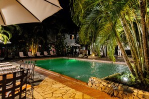 Un área de piscina nocturna con sillones y sombrillas, rodeada de palmeras y senderos de piedra.