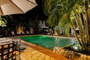 Un área de piscina nocturna con sillones y sombrillas, rodeada de palmeras y senderos de piedra.