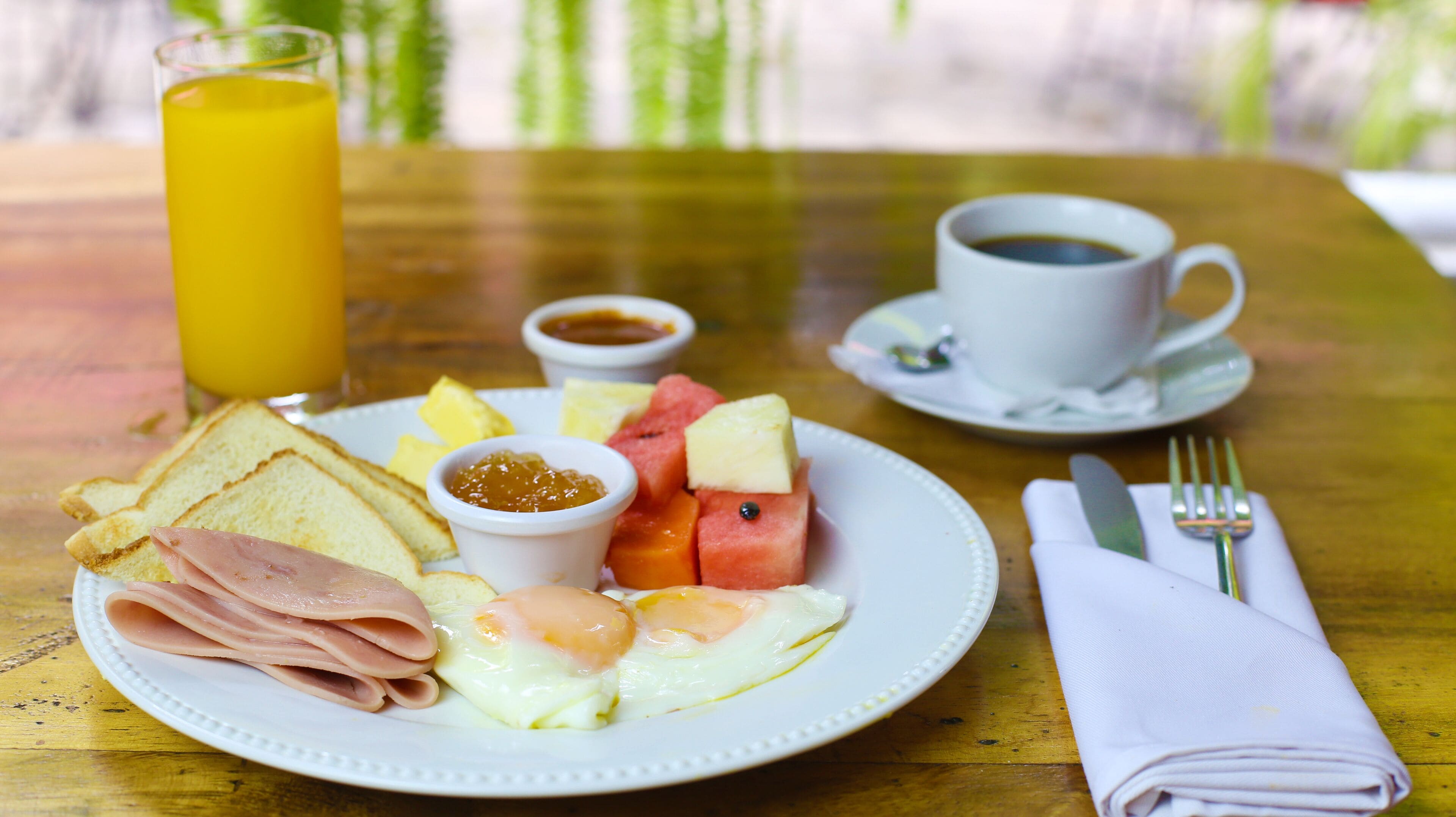 A breakfast plate with ham, eggs, toast, and fruit, a glass of orange juice, and a cup of coffee on a wooden table.