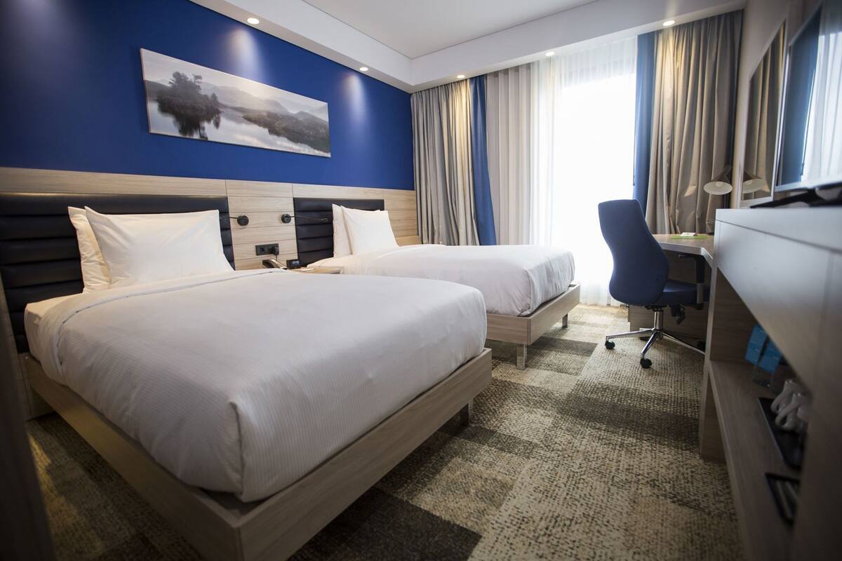 A hotel room with two beds, a desk, a chair, and a large window with curtains.