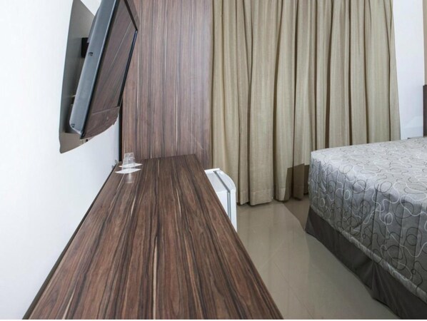 Una habitación de hotel con un escritorio de madera, una cama con ropa de cama estampada, un televisor montado y una ventana con cortinas.