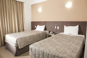 Quarto de hotel com duas camas, colchas bege estampadas, cabeceira de madeira e cortina à esquerda.