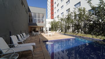 A modern outdoor pool area with lounge chairs and a table, surrounded by a building with multiple windows and a red accent wall.