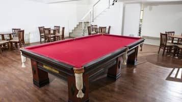 Uma mesa de bilhar com feltro vermelho, rodeada por cadeiras e mesas de madeira em um espaço com piso de madeira e paredes brancas.