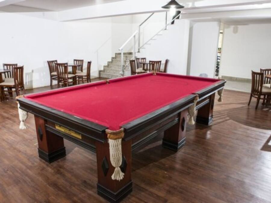 Una mesa de billar con superficie de fieltro rojo, rodeada de sillas y mesas de madera en una habitación con suelo de madera y paredes blancas.
