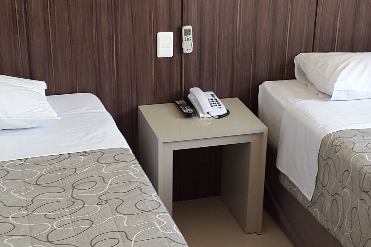 Une chambre d’hôtel avec deux lits, chacun agrémenté d’un oreiller blanc et d’une couvre-lit à motifs. Une petite table avec un téléphone et une télécommande se trouve entre les deux lits.