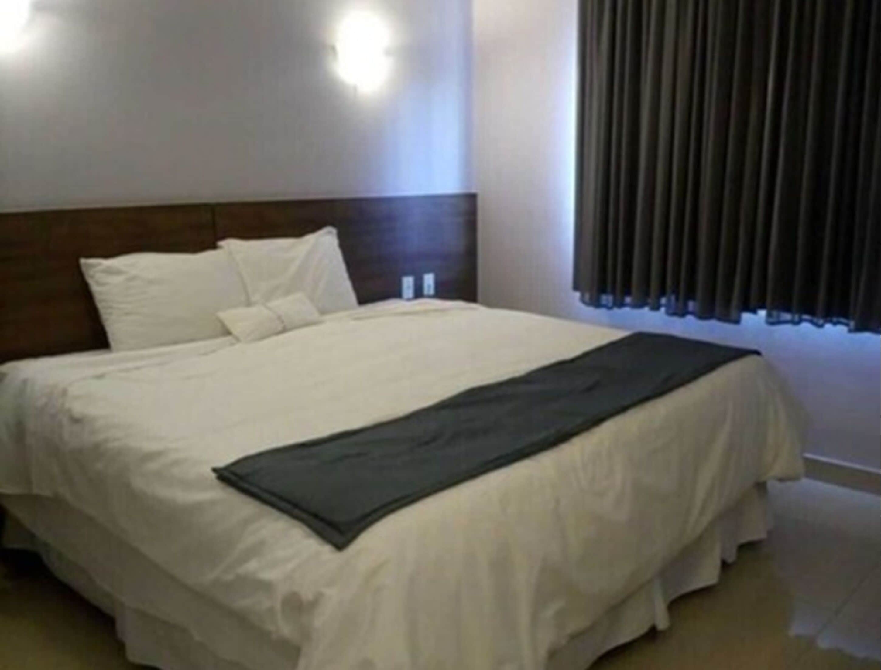 A hotel room with a large bed, white bedding, a dark headboard, and a window with curtains.