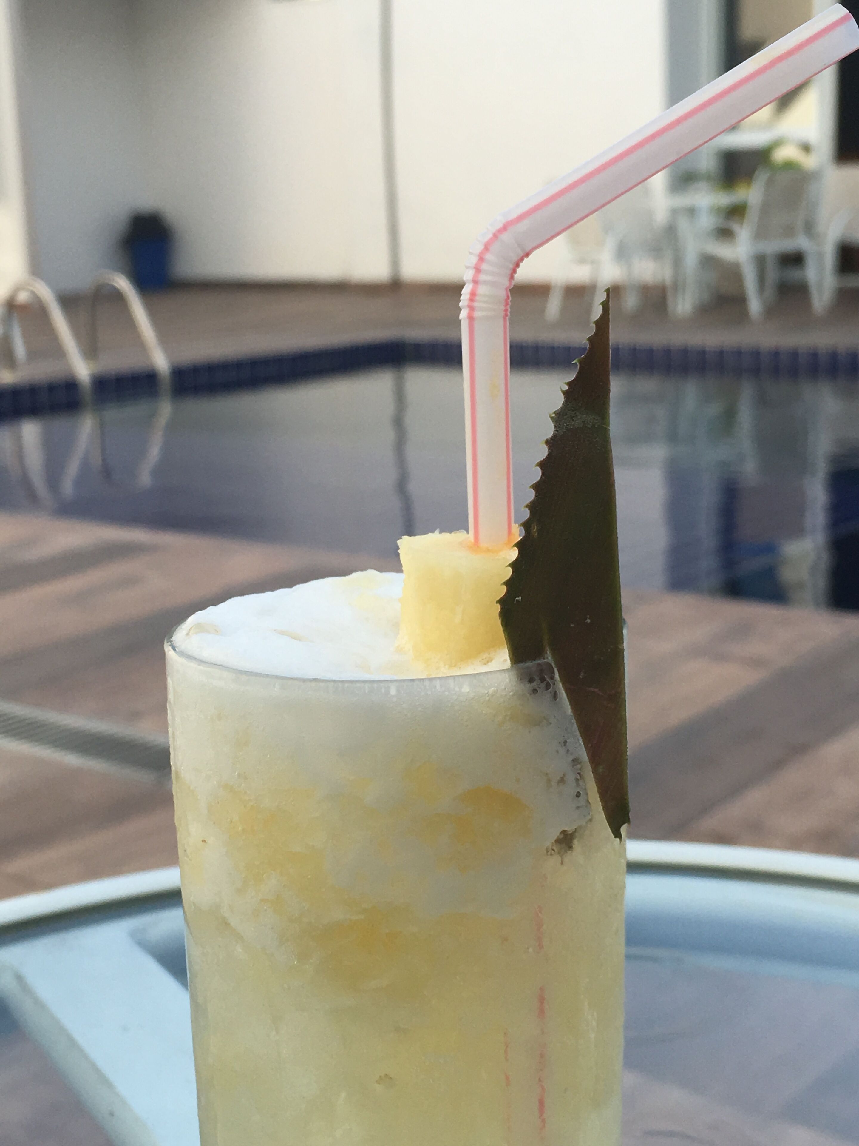Um copo com canudo e um pedaço de abacaxi sobre uma mesa perto de uma piscina.