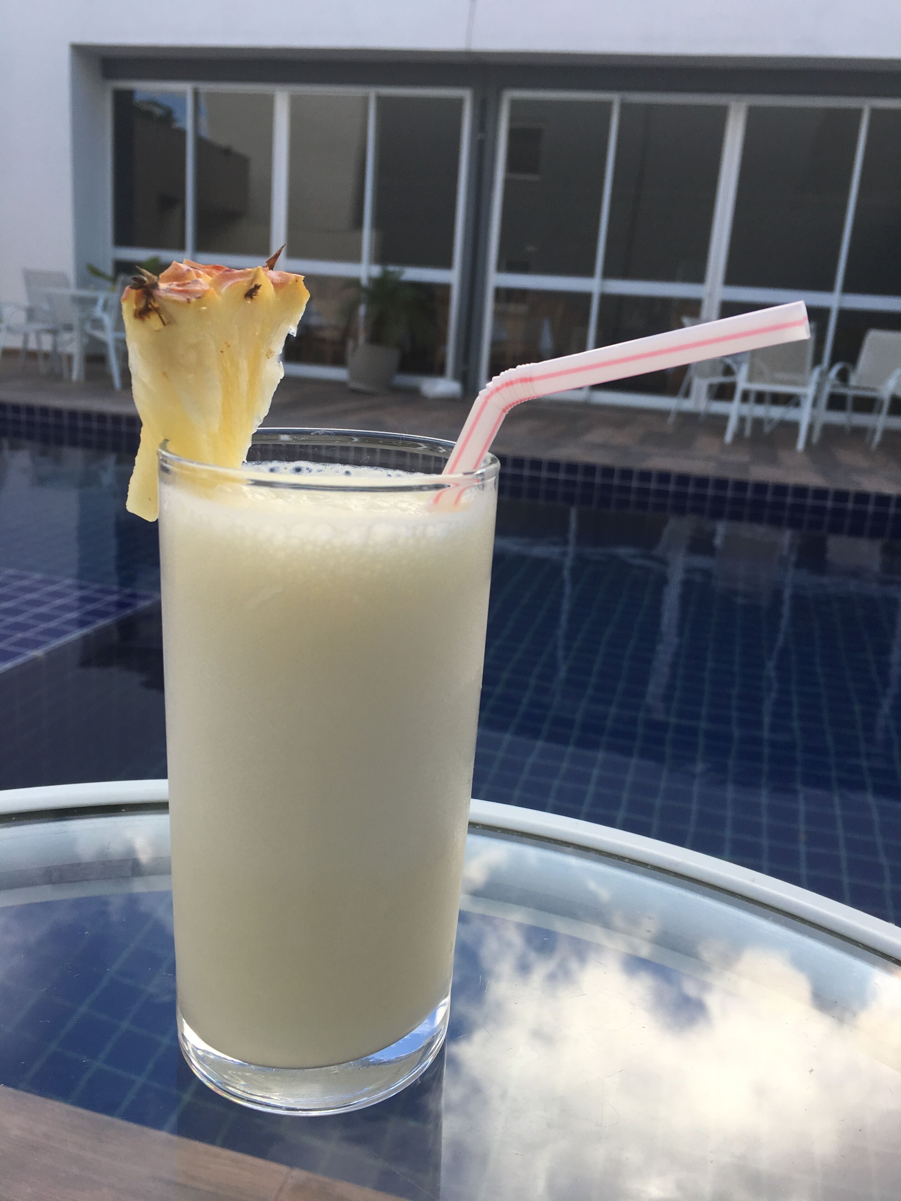 Ein Glas helles Getränk mit Trinkhalm und Ananasstück am Rand, auf einem Glastisch in Poolnähe.