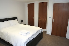 A neatly made bed with white linens and a folded towel, a bedside lamp, and three wooden doors in a bedroom.