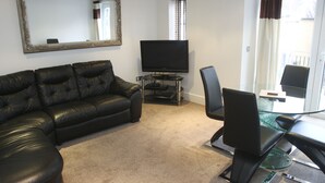 A living room with a black leather sofa, a flat-screen TV on a stand, a glass coffee table, and a black office chair. - 2 Bed Apt in Chorleywood Near Station (Rickmansworth)