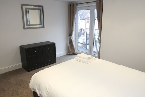 A bedroom with a bed, a dresser, a window with curtains, and a view of buildings outside.