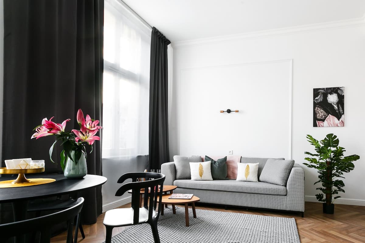 Ein modernes Wohnzimmer mit einem schwarz-weiß karierten Sofa, einem Esstisch mit Stühlen, einem Couchtisch, einem schwarz-weiß gemusterten Teppich, einer Topfpflanze und einer Küchenzeile mit Schränken und einem Wasserkocher.