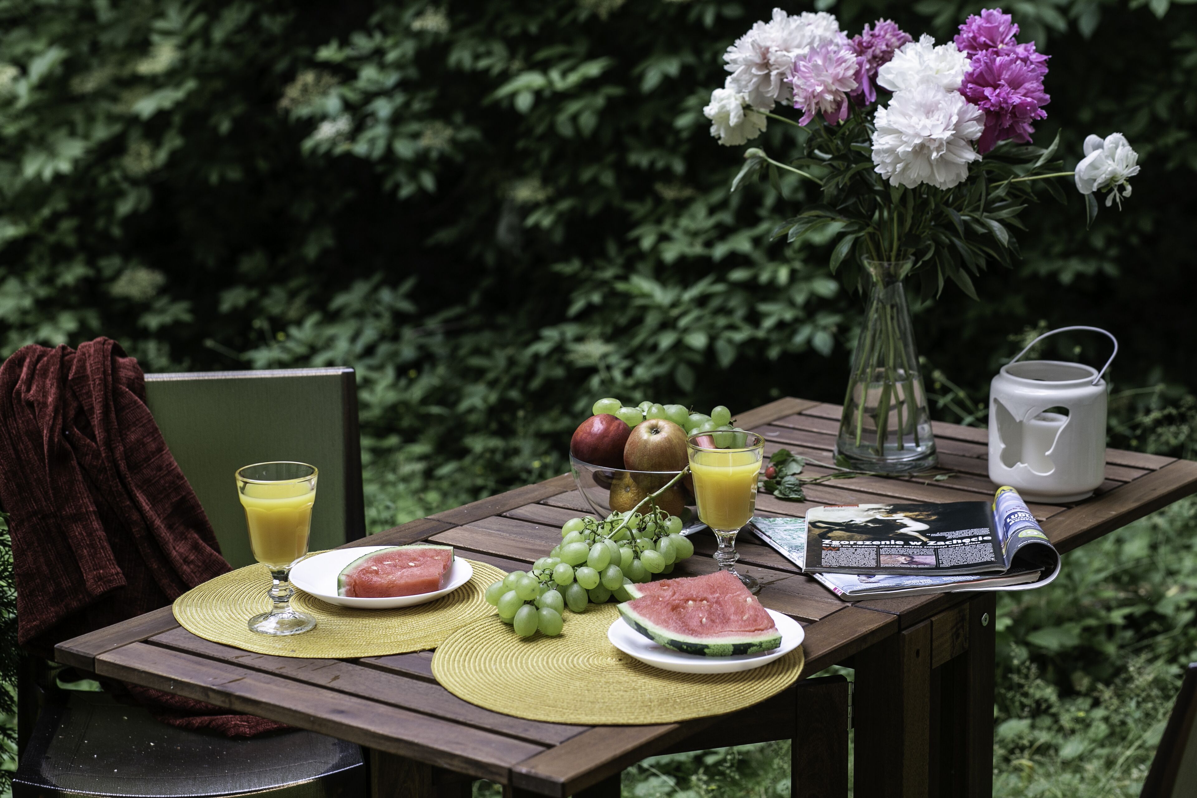Una mesa de madera con un mantel individual, un vaso de jugo de naranja, un plato con sandía y uvas, un tazón de fruta y un florero con flores.