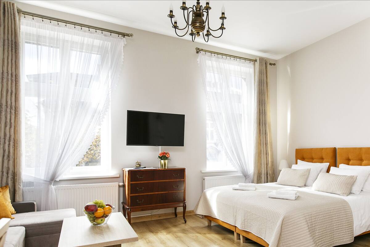 Ein Hotelzimmer mit einem Bett, einem Fernseher, einem Schminktisch, einem Schreibtisch mit Obstschale und einem Fenster mit Vorhängen.