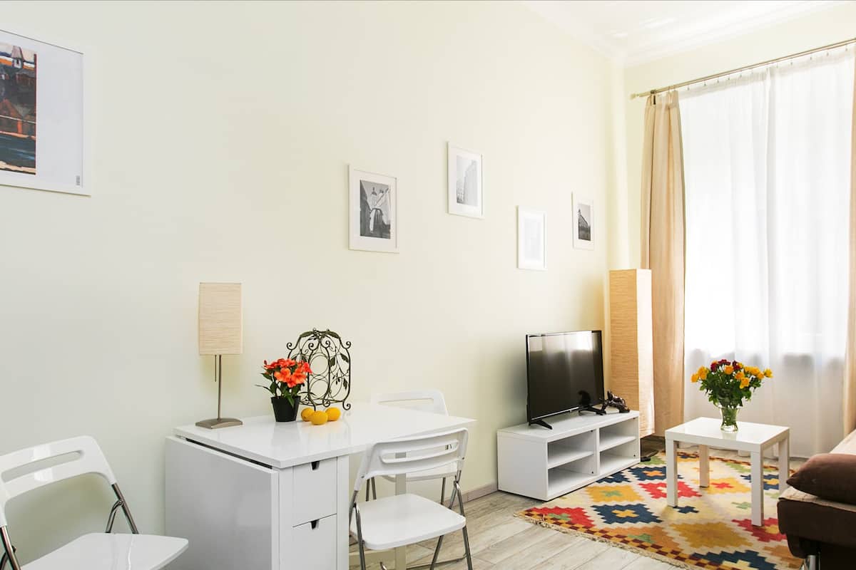 Ein Wohnzimmer mit einem weißen Schreibtisch, einem Fernseher, einem braunen Sofa und einem farbenfrohen Teppich.