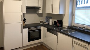 A compact kitchen with white cabinets, a black countertop, a built-in oven, a dishwasher, and a window with blinds. - Ferienpark Thüle (Friesoythe)