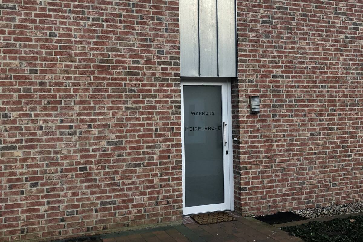 Un bâtiment en briques avec une porte blanche portant l’inscription « Wohnung Heidelederche » et deux fenêtres équipées de stores.