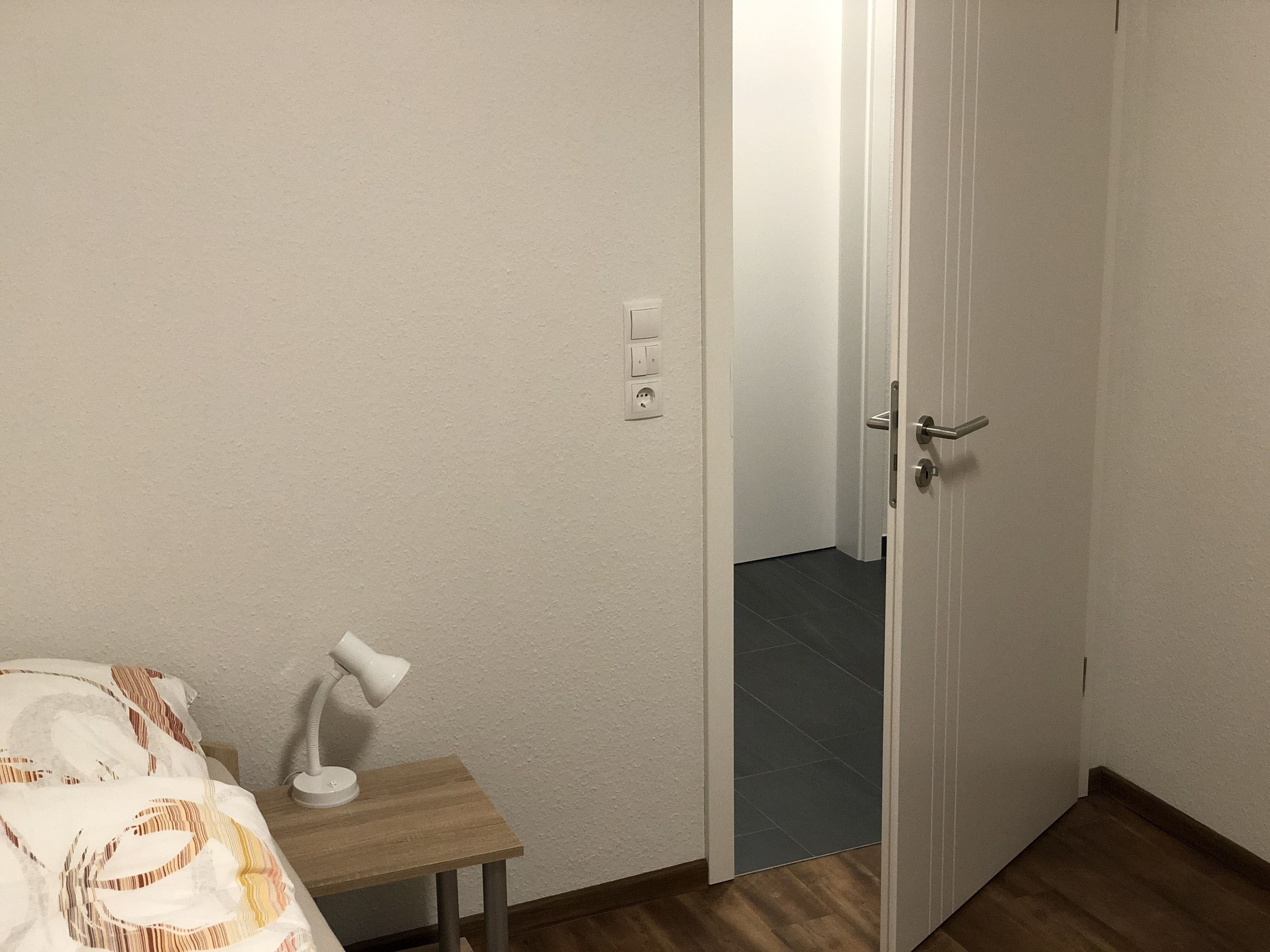 Un dormitorio con cama, mesita de noche, lámpara y una puerta que da a otra habitación.