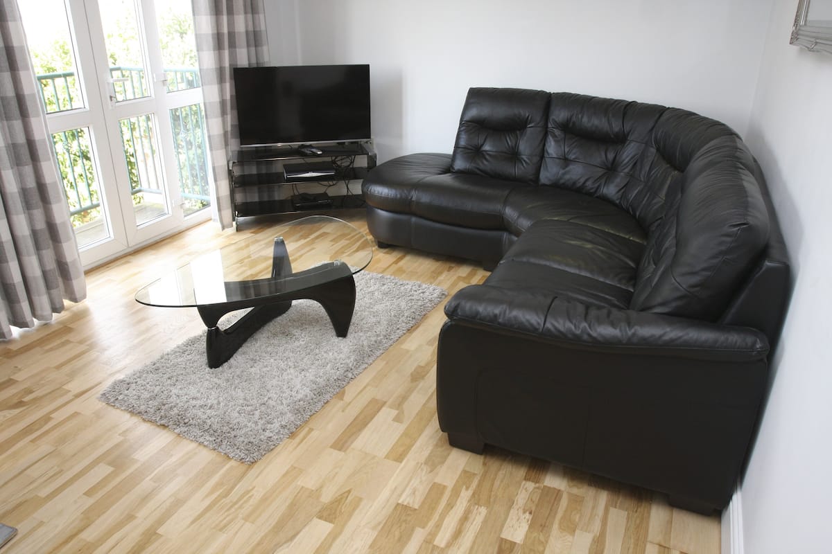 A modern living room with a black leather sofa, a glass coffee table, and a flat-screen TV on a stand.