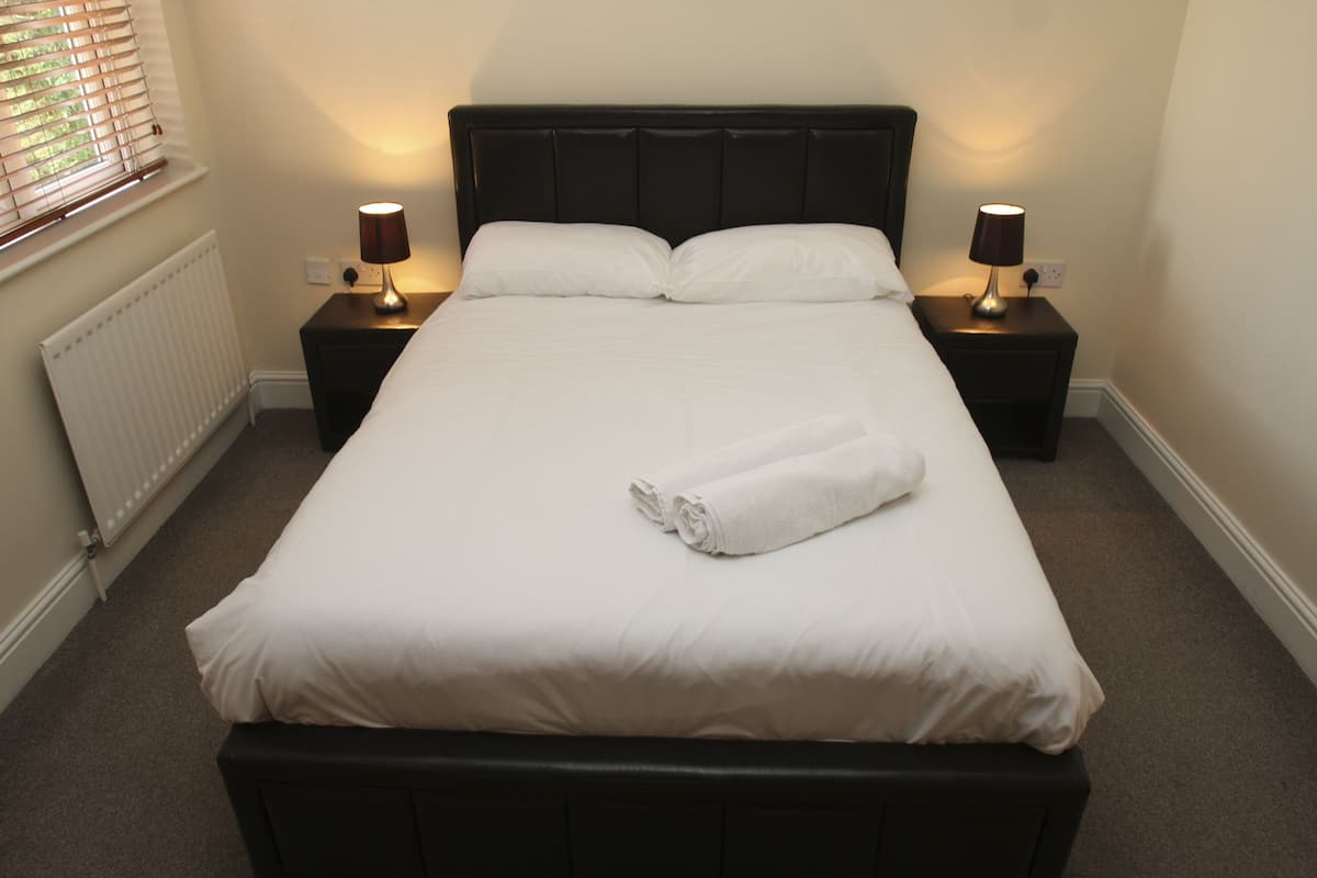 A neatly made bed with white linens and a rolled towel, flanked by two bedside tables with lamps, in a room with a radiator and a window with blinds.