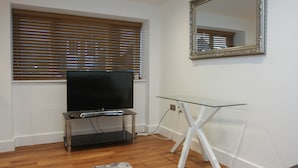 A modern living room with a flat-screen TV on a glass table, wooden flooring, and a large mirror on the wall.