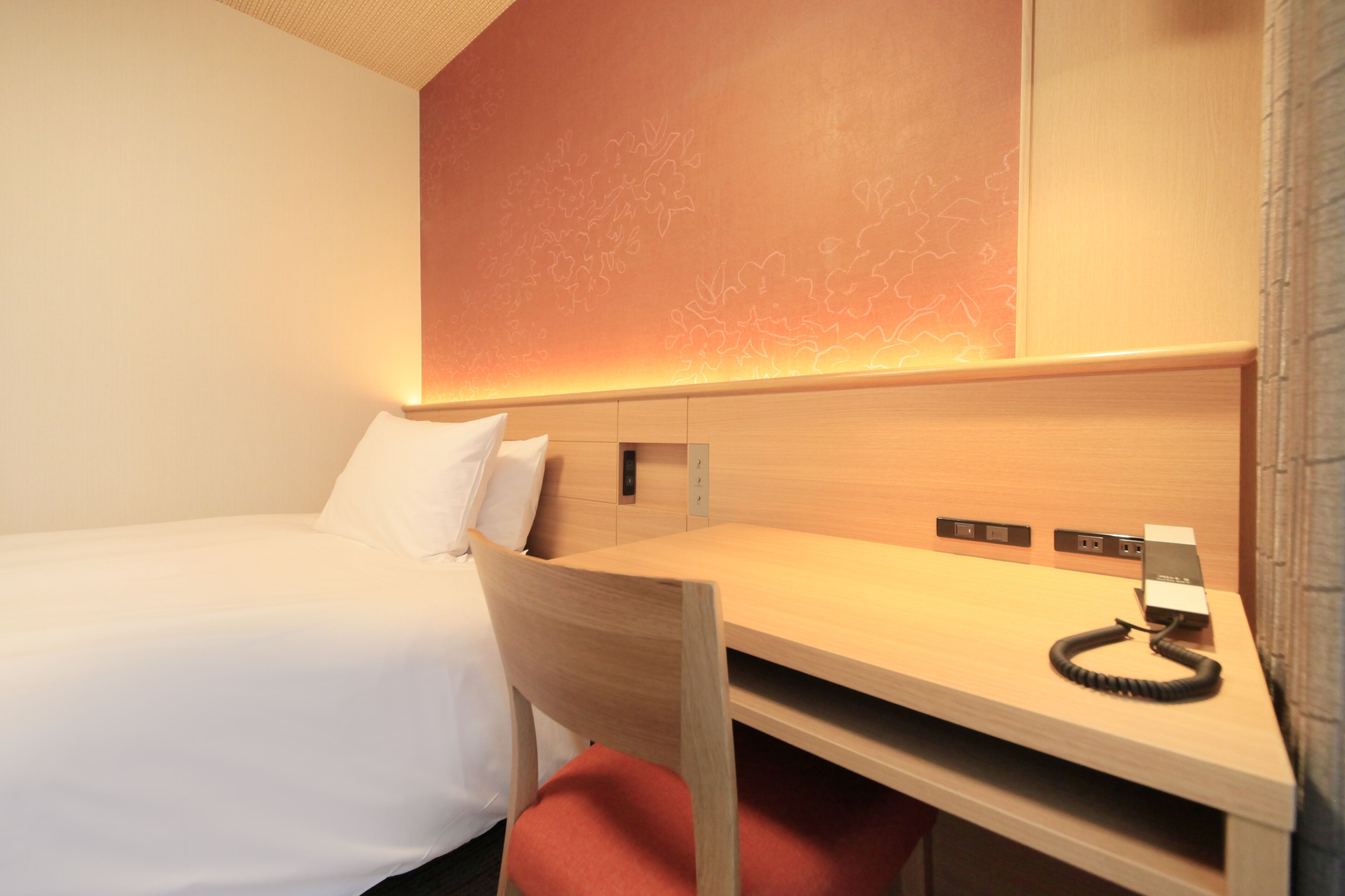 A hotel room with a single bed, a desk with a telephone, and a wall-mounted control panel.