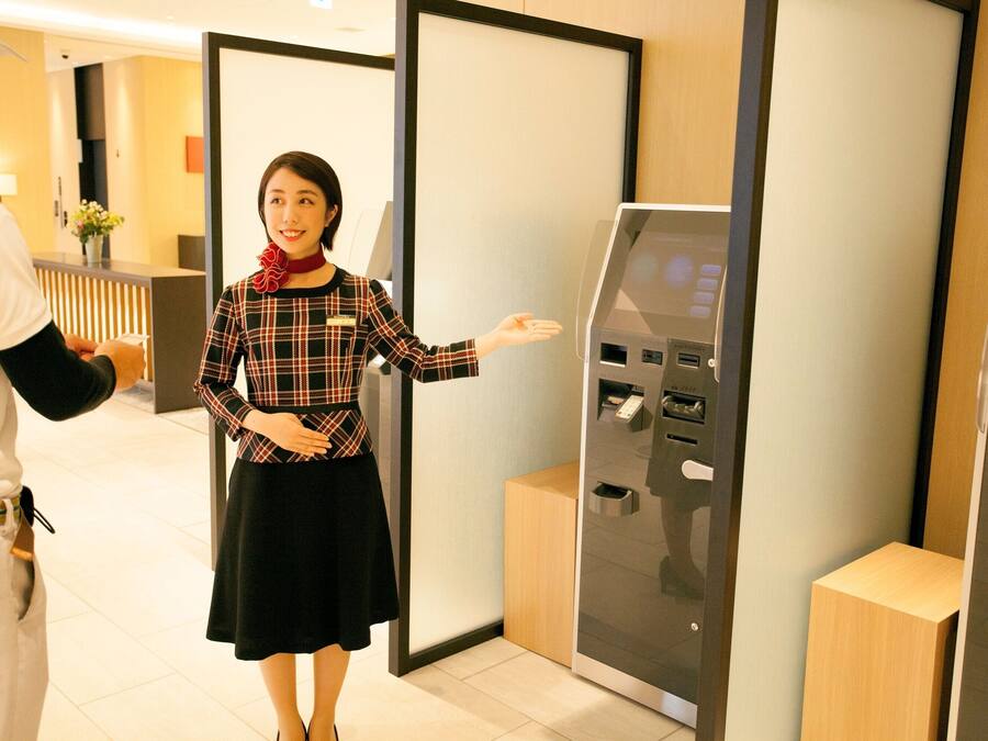 A hotel reception area with a modern design, featuring a reception desk, a self-service kiosk, and a wooden accent wall.