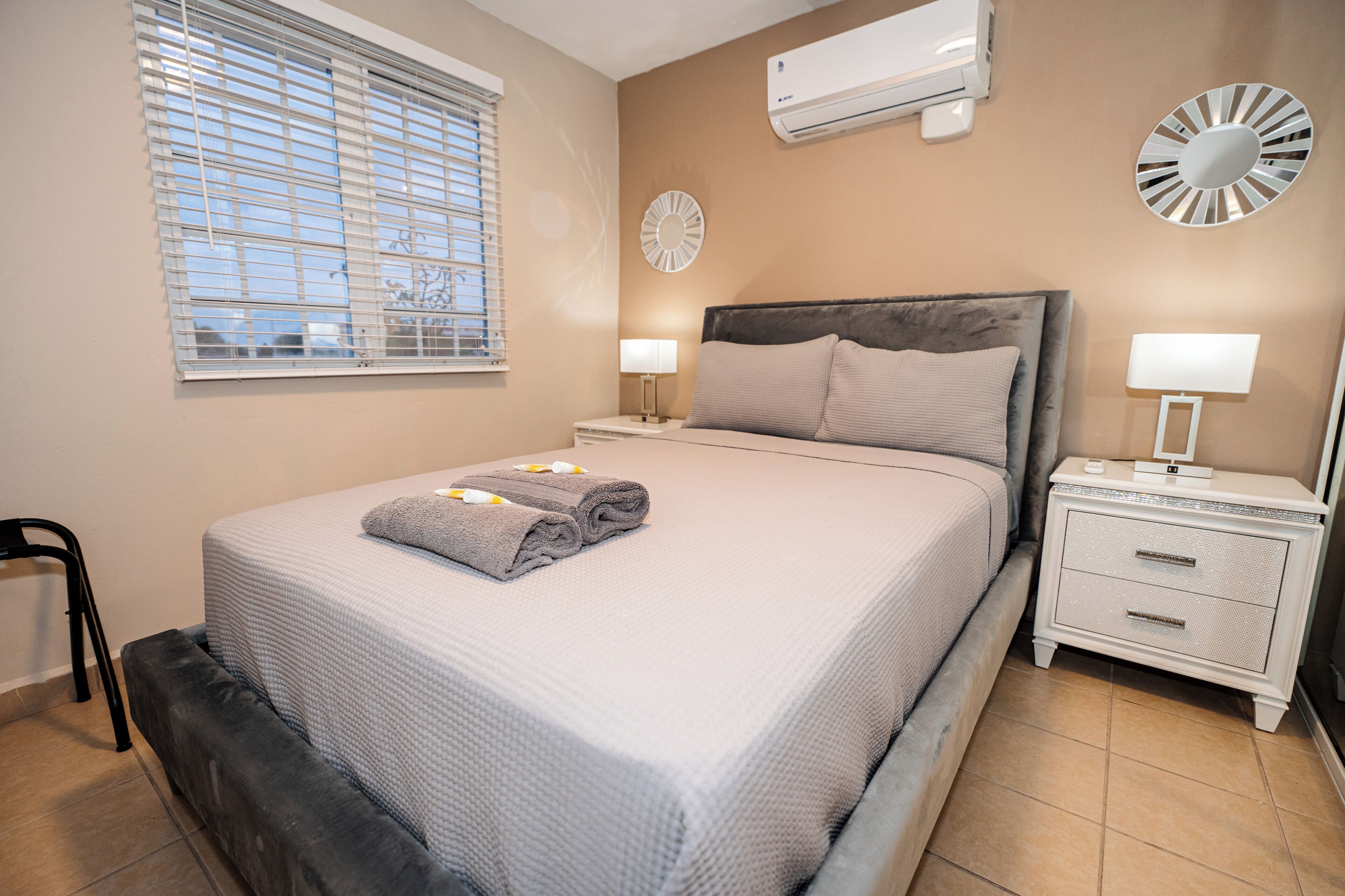 A neatly made bed with a grey blanket and a folded towel, a bedside table with a lamp, and a wall-mounted air conditioner.