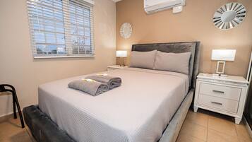 A neatly made bed with a grey blanket and a folded towel, a bedside table with a lamp, and a wall-mounted air conditioner.