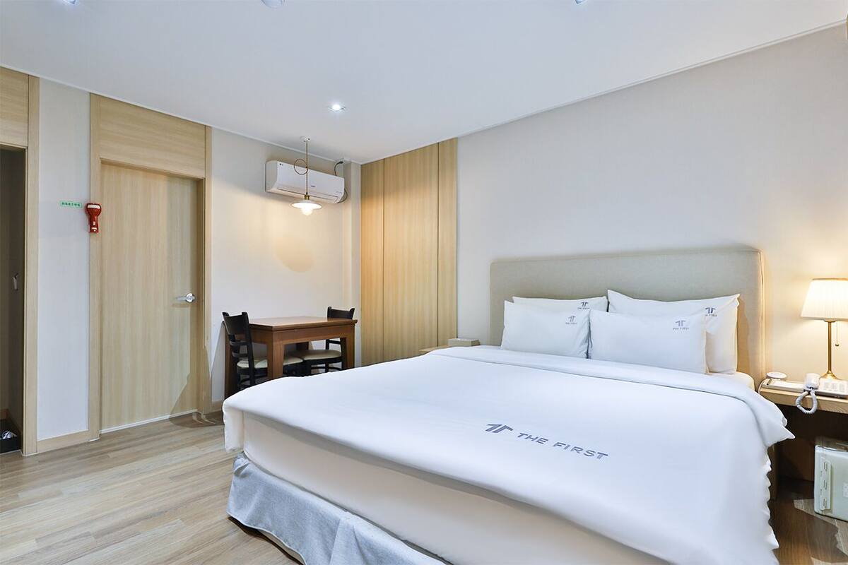 Ein Hotelzimmer mit Bett, Schreibtisch und Stuhl. Eine Tür und ein Klimagerät sind an der Wand angebracht.