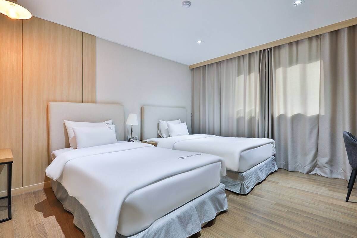 Ein Hotelzimmer mit zwei Betten, einem Schreibtisch, einem Stuhl und einem großen Fenster mit Vorhängen.