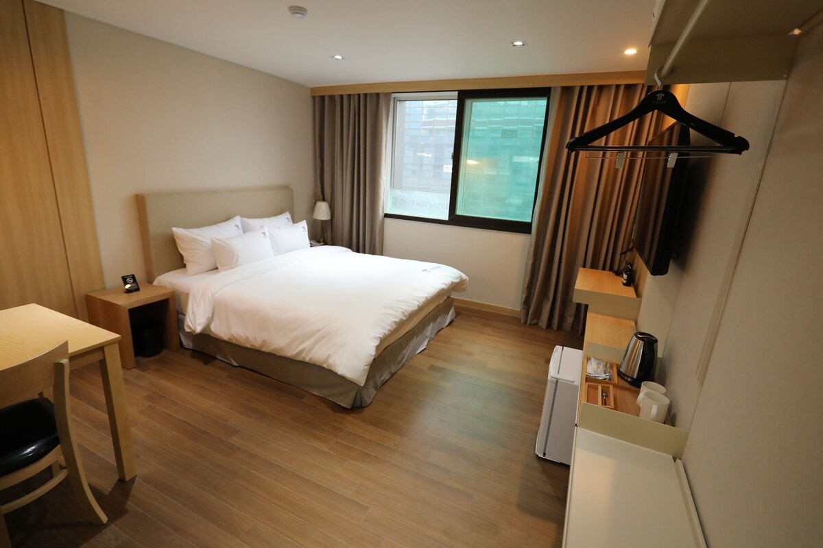 A hotel room with a bed, bedside table, lamp, window, and a coat rack.