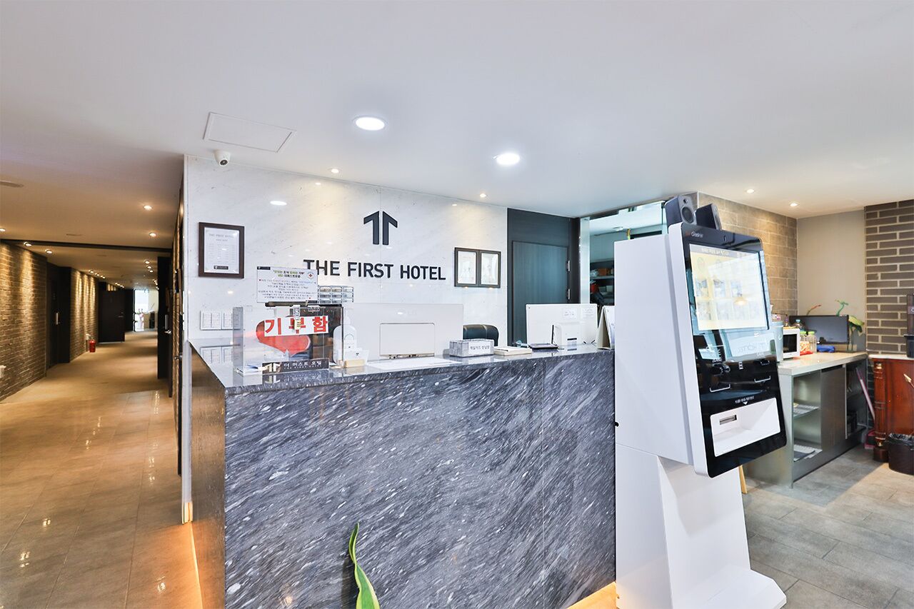 Zona de recepción de hotel con un mostrador de mármol, un quiosco de información digital y un cartel con el nombre del hotel.