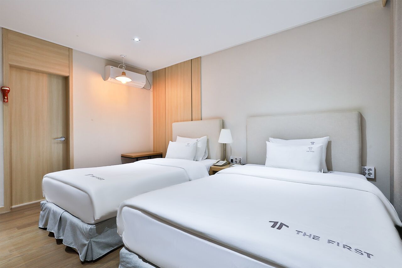 A hotel room with two beds, a bedside table with a lamp, a wall-mounted air conditioner, and a door with a red handle.