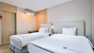 A hotel room with two beds, a bedside table with a lamp, a wall-mounted air conditioner, and a door with a red handle.
