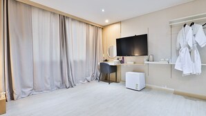 Una habitación de hotel moderna con un ventanal grande, un televisor de pantalla plana, un escritorio con una silla, un refrigerador pequeño y un perchero con batas.