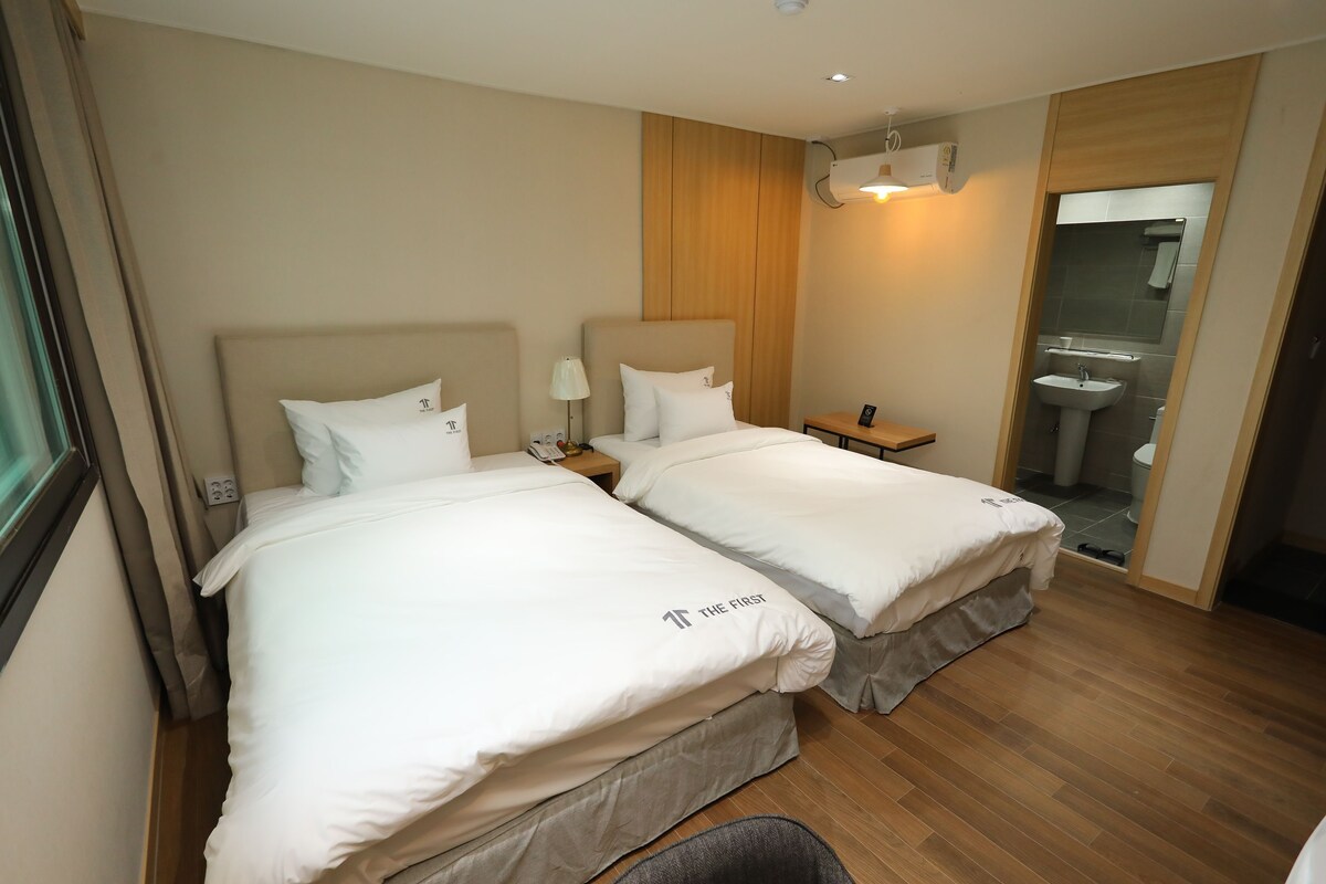 A hotel room with two beds, a bedside table with a lamp, a wooden headboard, a small desk, and a bathroom visible through an open door.