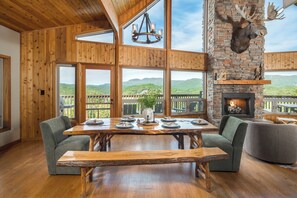 Interior de madera con una gran mesa de comedor, asientos acolchados, chimenea y vistas a la montaña.