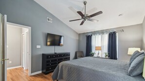 Ein Schlafzimmer mit Bett, Deckenventilator, Fernseher, Kommode und einem Fenster mit Vorhängen.