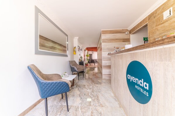 A reception area with a blue sign for Ayenda Hotels, a counter with a mirror, and seating with a blue chair and a small table.
