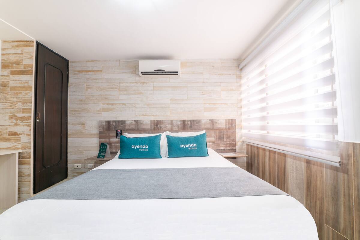 A hotel room with a bed, two pillows, a nightstand, a wall-mounted air conditioner, and a window with blinds.