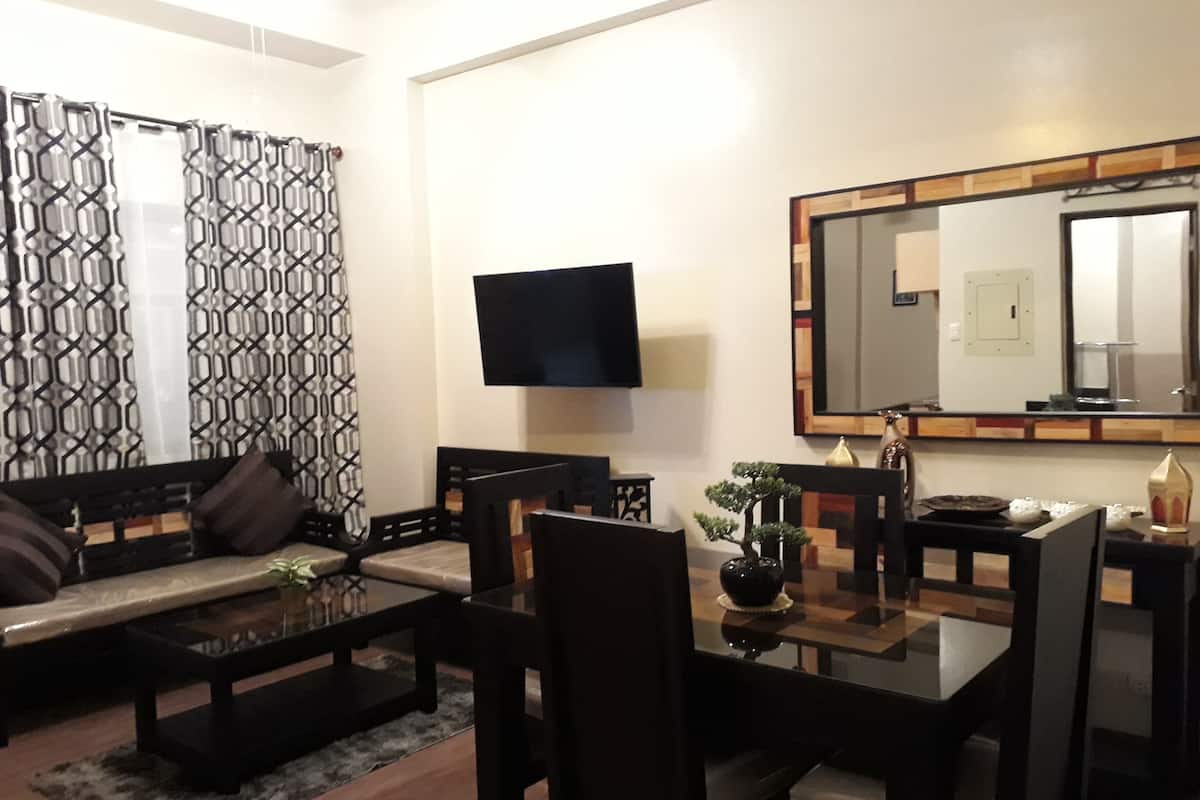 A modern living room with a sofa, coffee table, and dining table. A flat-screen TV is mounted on the wall, and a large mirror is above the dining table.