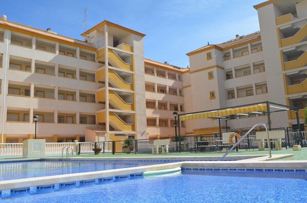 A multi-story hotel with a swimming pool, balconies, and a covered area with seating.