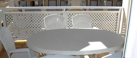 A balcony with a round table and white plastic chairs, overlooking a residential area with buildings and a mountain in the background.