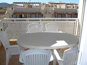 A balcony with a round table and white plastic chairs, overlooking a residential area with buildings and a mountain in the background.
