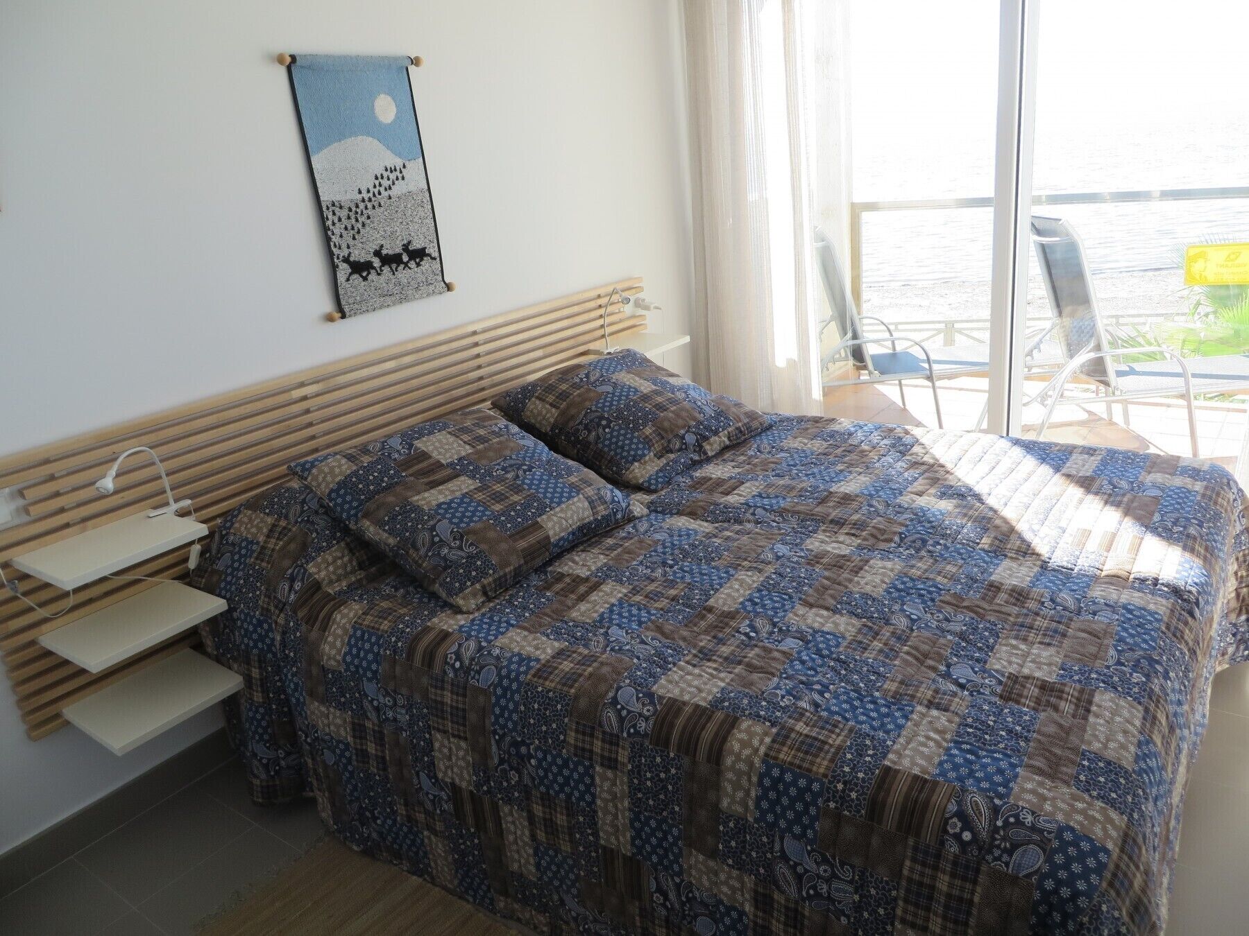 A bedroom with a bed, a bedside table, a wall hanging, and a view of the outdoors.