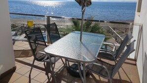 Een balkon met een glazen tafel en stoelen, met uitzicht op een strand en de zee.
