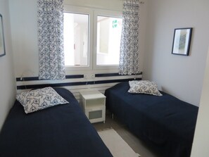A small, clean room with two single beds, a window with curtains, a bedside table, and a framed picture on the wall. - 1st floor sea view apartment, free wifi, communal pool (La Manga del Mar Menor)